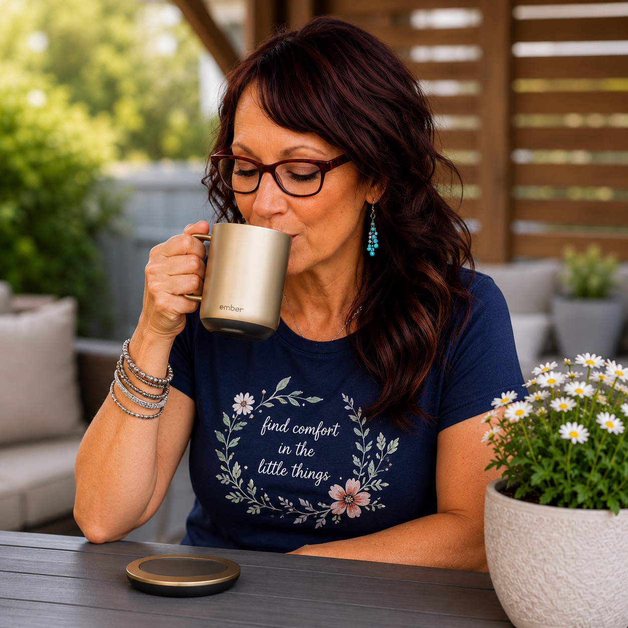 Woman with dark shoulder-length hair and glasses enjoying a drink from a gold Ember Smart Temperature Control Mug while seated at an outdoor patio table. The mug's charging coaster sits on the table beside a potted daisy plant in a cozy backyard setting.